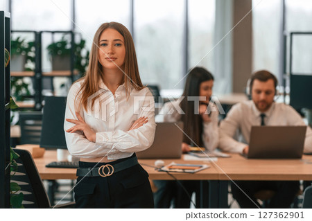 Woman with arms crossed is standing. Business people in formal clothes are working in the office together Woman with arms crossed is standing. Business people in formal clothes are working in the office together 127362901
