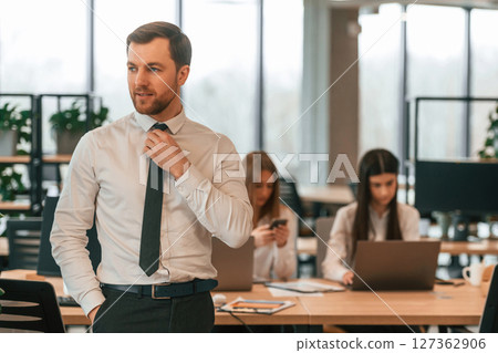 Man in tie is standing in front of his colleagues. Business people in formal clothes are working in the office together Man in tie is standing in front of his colleagues. Business people in formal clothes are working in the office together 127362906