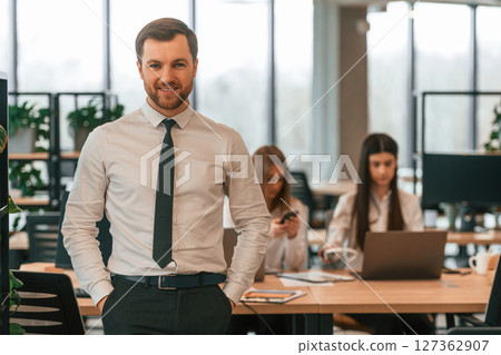 Man in tie is standing in front of his colleagues. Business people in formal clothes are working in the office together Man in tie is standing in front of his colleagues. Business people in formal clothes are working in the office together 127362907