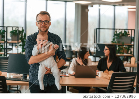 Man in glasses is standing and holding his son. Infant baby is in the office where group of people are working together 127362996