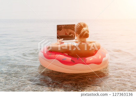 Woman Laptop Beach - Working Remotely on Vacation, Sitting in Inflatable Ring on Water, Sun Setting in the Background. 127363136