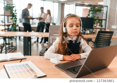 Sitting by the table with laptop. Little girl is in the office where people working together Sitting by the table with laptop. Little girl is in the office where people working together 127363861