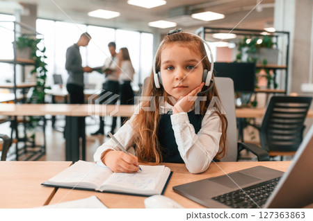Sitting by the table with laptop. Little girl is in the office where people working together Sitting by the table with laptop. Little girl is in the office where people working together 127363863