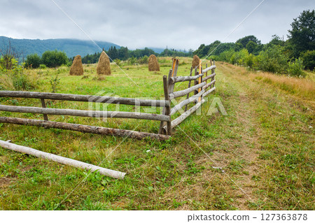 rural field behind the wooden fence. countryside landscape in mountains on a rainy day. organic haystack on the meadow 127363878