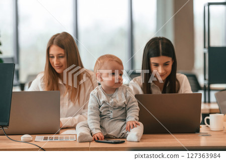 Sitting on the table. Women are using laptops. Infant baby is in the office where group of people are working together 127363984
