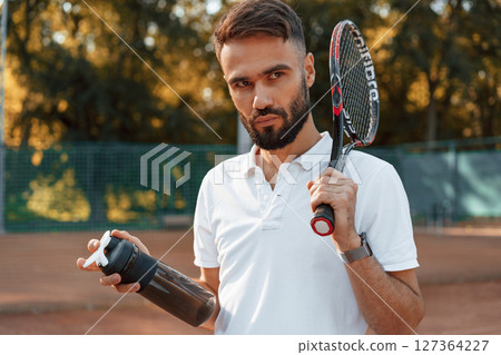 Holding bottle with water and racket. Young man is on the tennis court at sunny daytime 127364227