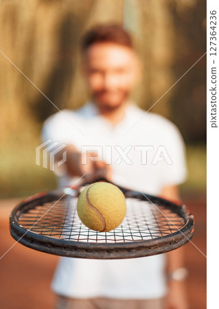 Close up view of racket and ball on it. Young man is on the tennis court at sunny daytime Close up view of racket and ball on it. Young man is on the tennis court at sunny daytime 127364236