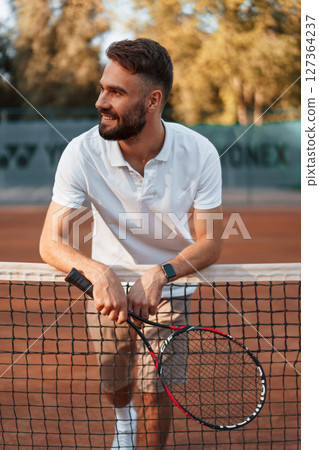 Standing by the net with racket. Young man is on the tennis court at sunny daytime 127364237