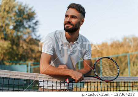 Standing with racket in hands. Young man is on the tennis court at sunny daytime 127364238