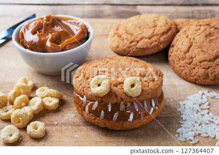 Cookies with cream paste in the shape of monsters for Halloween celebration. Funny homemade faces made of oatmeal cookies and boiled condensed milk. 127364407