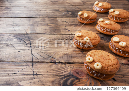 Cookies with cream paste in the shape of monsters for Halloween celebration. Funny homemade faces made of oatmeal cookies and boiled condensed milk. copy space 127364409