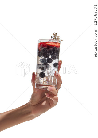 Female hand holding tall glass of fizzy drink with floating berries, ice cubes, and tiny flowers. Female hand holding tall glass of fizzy drink with floating berries, ice cubes, and tiny flowers. 127365371