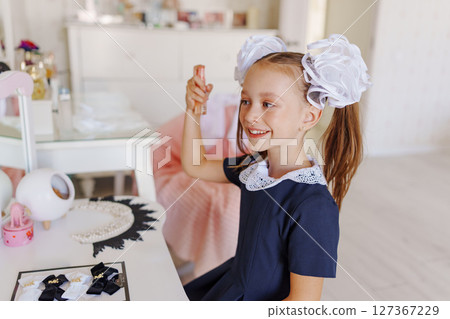 A little girl in a school uniform preens herself at a dressing table.  127367229