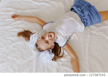 Cheerful little girl with ponytails and white bows lying on the bed. 127367243