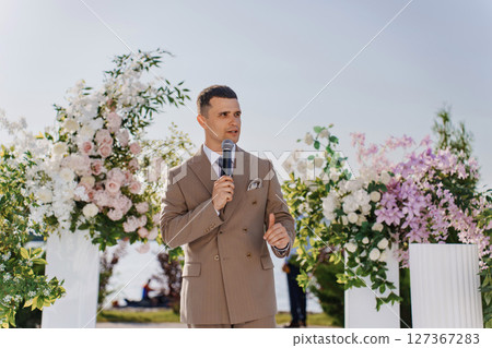 A man presenter in a beige suit leads a wedding ceremony in the park. A man presenter in a beige suit leads a wedding ceremony in the park. 127367283