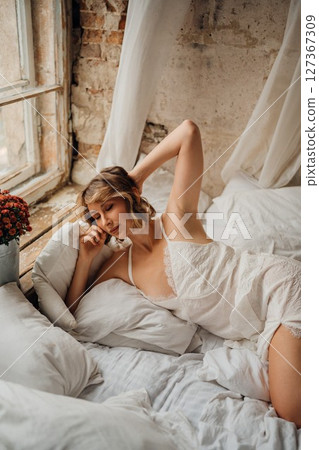 a blonde woman in a white dress on white sheets by the window 127367309