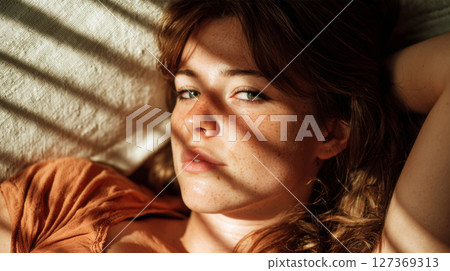 Young woman with freckles relaxing on a couch, sunlight casting shadows across her face, capturing a moment of serene introspection and natural beauty 127369313