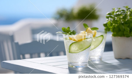 Two spa spritz cocktails with cucumber and lemon sit on a white table under a clear blue sky Two spa spritz cocktails with cucumber and lemon sit on a white table under a clear blue sky 127369374