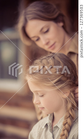 Cowgirl mother lovingly braids her daughter's hair on a rustic porch, capturing a beautiful family moment 127369583