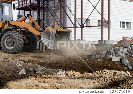 A powerful front loader on a construction site clears up the battle after dismantling reinforced concrete structures. Moving soil with construction equipment. Excavations. A powerful front loader on a construction site clears up the battle after dismantling reinforced concrete structures. Moving soil with construction equipment. Excavations. 127371619
