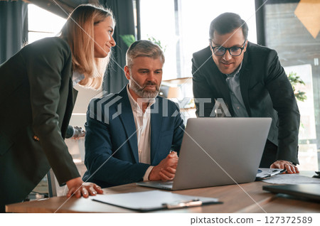 Senior man is sitting by laptop, employees are helping. Group of business people are working in the modern office Senior man is sitting by laptop, employees are helping. Group of business people are working in the modern office 127372589