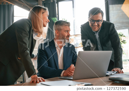 Senior man is sitting by laptop, employees are helping. Group of business people are working in the modern office 127372590