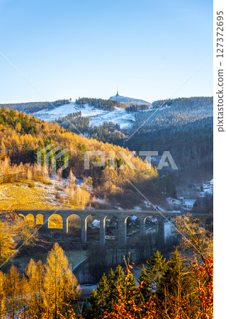 A stone railway bridge in Novina spans across a snowy valley, with the Jested Mountain rising majestically in the background during a picturesque winter sunset. A stone railway bridge in Novina spans across a snowy valley, with the Jested Mountain rising majestically in the background during a picturesque winter sunset. 127372695