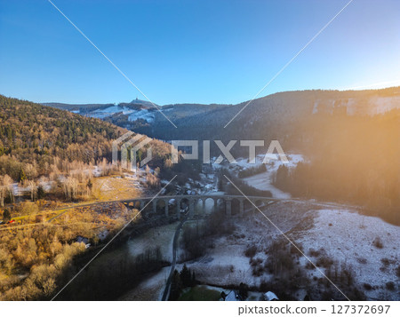 A striking railway stone bridge stands in Novina, Krystofovo udoli, embraced by snow-covered hills. Jested Mountain rises majestically in the background during a serene winter sunset. A striking railway stone bridge stands in Novina, Krystofovo udoli, embraced by snow-covered hills. Jested Mountain rises majestically in the background during a serene winter sunset. 127372697
