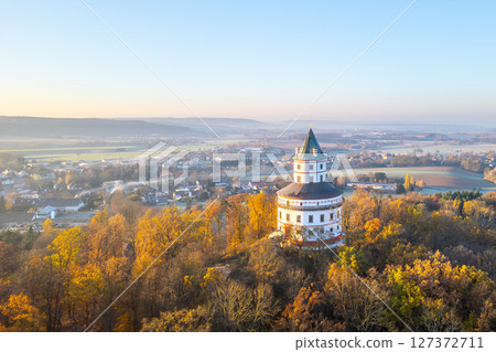 The elegant Baroque hunting chateau Humprecht stands above Sobotka, surrounded by vibrant autumn foliage, illuminated by the soft light of a tranquil sunrise. 127372711