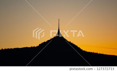 The silhouette of Jested Tower rises against a vibrant evening sky in Liberec, Czechia. The last light of day casts a warm glow, highlighting the tower's distinctive shape. The silhouette of Jested Tower rises against a vibrant evening sky in Liberec, Czechia. The last light of day casts a warm glow, highlighting the tower's distinctive shape. 127372713