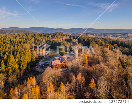 Bramberk lookout tower rises above the colorful autumn landscape of the Jizera Mountains. Sunlight bathes the area, highlighting the beauty of the surrounding trees and peaceful atmosphere. 127372714