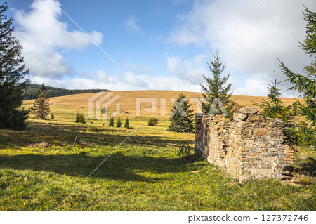 The remnants of the Konigsmuhle settlement display weathered stone walls surrounded by lush greenery and rolling hills in the Ore Mountains of Czechia on a sunny day. 127372746