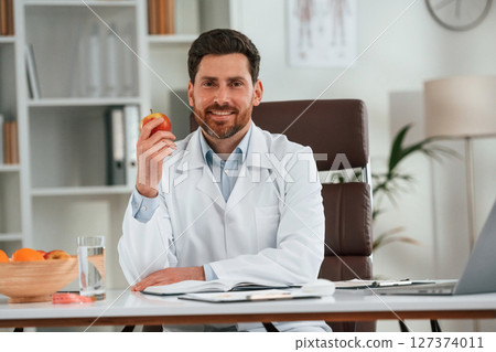Holding an apple. Doctor in formal clothes is working in the cabinet Holding an apple. Doctor in formal clothes is working in the cabinet 127374011