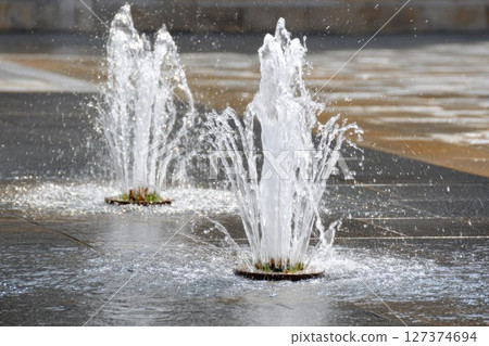 Water fountains spraying in a modern urban plaza with smooth stone pavement and greenery 127374694