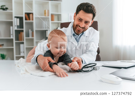Toddler is lying down on the table. Doctor with little baby is working in the cabinet Toddler is lying down on the table. Doctor with little baby is working in the cabinet 127374792