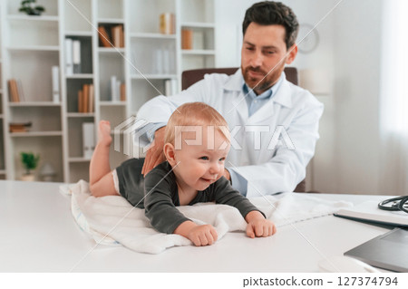 Toddler is lying down on the table. Doctor with little baby is working in the cabinet 127374794