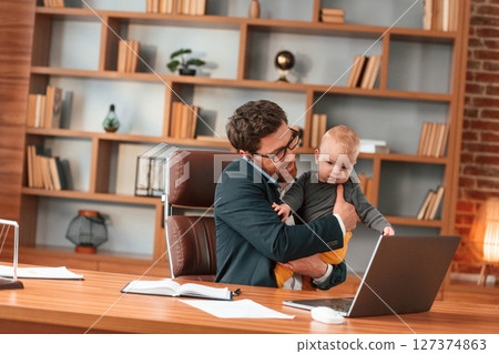 Conception of multitasks. Businessman in formal clothes is working indoors. With toddler 127374863
