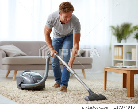 Man using vacuum cleaner to clean carpet in bright living room with modern furniture and decor 127374919