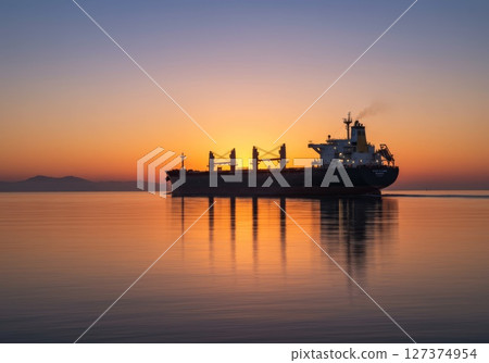 Cargo ship sailing on calm waters during sunset with vibrant colors reflecting on surface 127374954