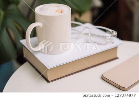 Cozy home setup with cappuccino, book, eyeglasses, and smartphone on a round table 127374977