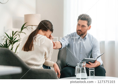 Woman is crying on the therapy session. Doctor in formal clothes is working in the cabinet Woman is crying on the therapy session. Doctor in formal clothes is working in the cabinet 127375103