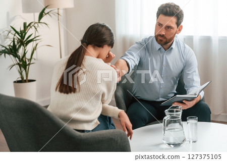 Woman is crying on the therapy session. Doctor in formal clothes is working in the cabinet Woman is crying on the therapy session. Doctor in formal clothes is working in the cabinet 127375105