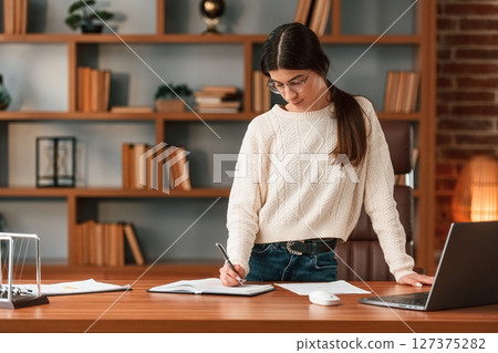 Young woman in glasses is working indoors. Standing by table with laptop Young woman in glasses is working indoors. Standing by table with laptop 127375282