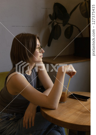young woman is sitting in a cafe with a glass of orange coffee and looking out the window. young woman is sitting in a cafe with a glass of orange coffee and looking out the window. 127375435