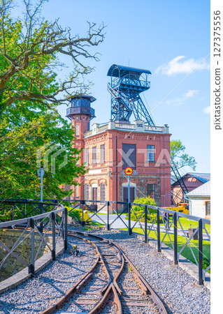 At Sevcinsky mine in Brezove hory, Pribram, Czechia, visitors explore a historical site with striking architecture and mining equipment surrounded by greenery and a clear blue sky. At Sevcinsky mine in Brezove hory, Pribram, Czechia, visitors explore a historical site with striking architecture and mining equipment surrounded by greenery and a clear blue sky. 127375556