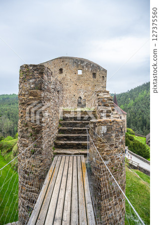 Visitors can walk along the wooden bridge leading to the historical ruins of Velhartice Castle in Czechia. Surrounded by lush greenery, it offers a glimpse into the past amidst cloudy skies. 127375560