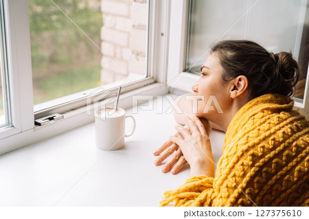 Woman relaxing with a cup of coffee near the window, wrapped in a yellow sweater 127375610