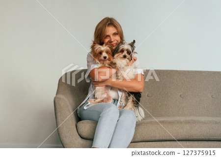 Holding and embracing the animals. Woman is sitting on a sofa with two cute dogs Holding and embracing the animals. Woman is sitting on a sofa with two cute dogs 127375915