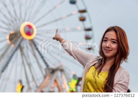 A joyful young woman pointing at a colorful ferris wheel, capturing the fun atmosphere of a carnival night. 127376012