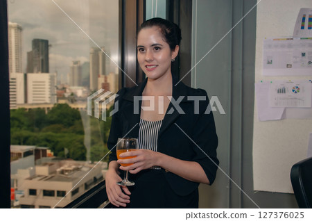 Professional woman holding a drink, dressed in a sleek outfit, with a city skyline backdrop, exuding confidence. Professional woman holding a drink, dressed in a sleek outfit, with a city skyline backdrop, exuding confidence. 127376025